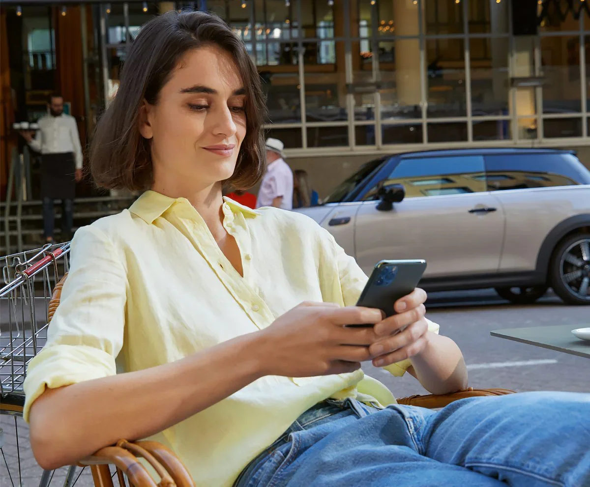Female wearing yellow collared button-down shirt and jeans, sitting in a chair and holding a smartphone next to a street with a grey MINI vehicle parked in the background. | MINI of Pittsburgh in Pittsburgh PA
