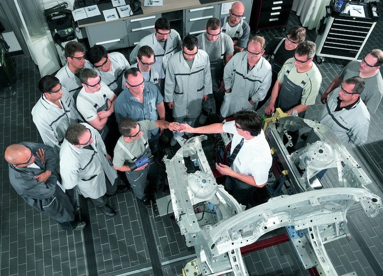 Overhead view of a group of men in safety glasses and workwear gathered around a disassembled car chassis.