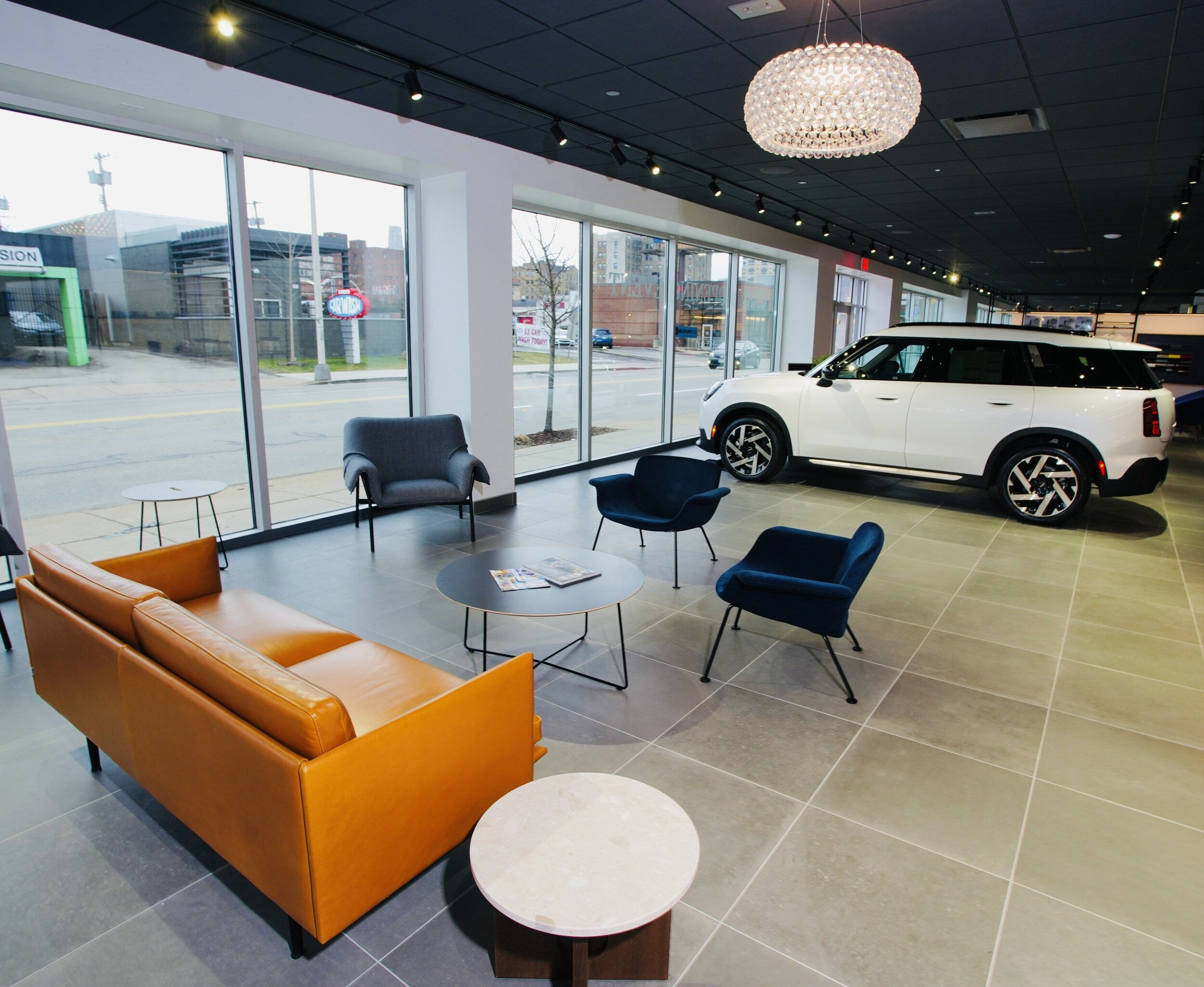 Modern car showroom with a white SUV, an orange couch, blue chairs, and a cityscape visible through large windows.