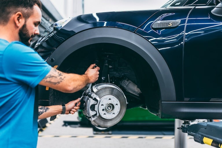 A mechanic works on the front brake of a dark blue car lifted in a garage.
