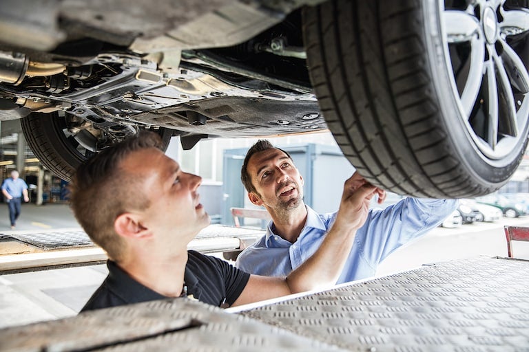 A mechanic and car owner inspect the underside of an elevated car.