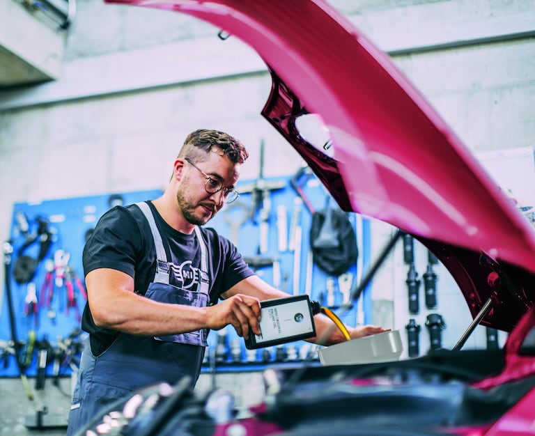 Mechanic wearing glasses and MINI branded overalls, pouring oil into a red car in a garage.