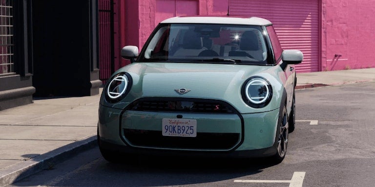 Front view of a light green Mini Cooper S with a white roof, parked on asphalt with a pink wall behind it.