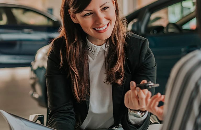 Smiling saleswoman with long brown hair, wearing a white shirt with a studded collar and a black blazer, holding out car keys.