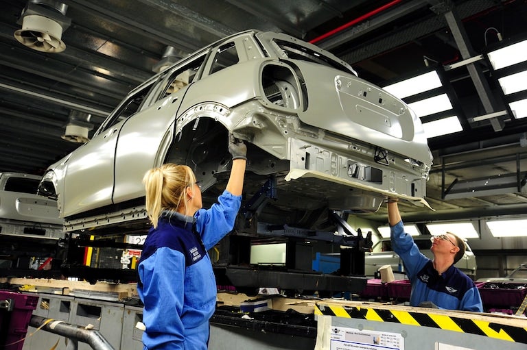 Two workers in blue uniforms assemble a car body frame on an assembly line.
