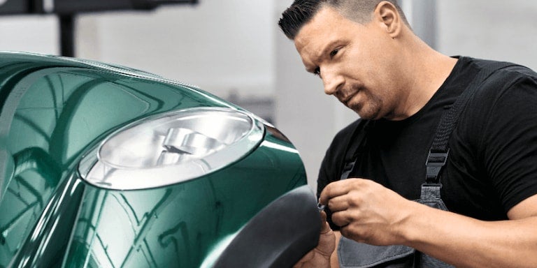 A male mechanic in black overalls works on the green front fender of a car.