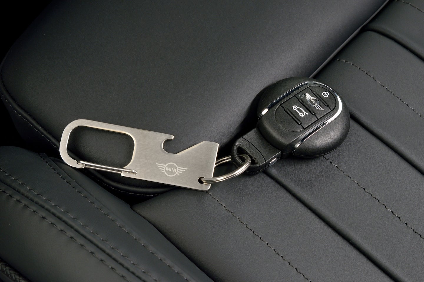 Mini Cooper car key fob and a metal carabiner bottle opener keychain with a Mini logo on a black leather seat.