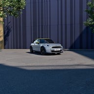 Silver Mini Cooper convertible parked on a concrete lot with a dark blue corrugated building and trees in the background.