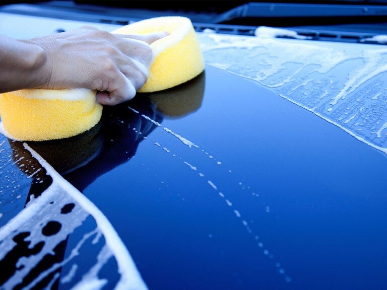 Hand in frame holding a yellow sponge, washing a dark blue car hood covered in soap suds.